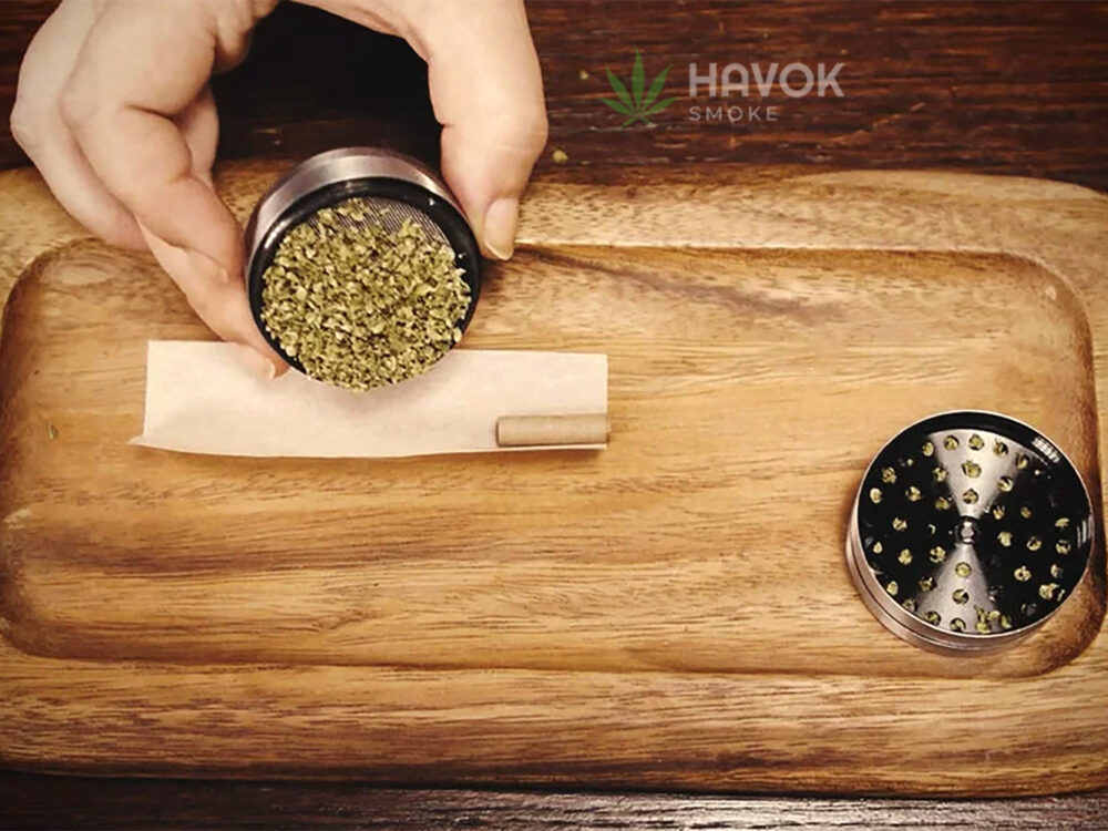 Hand holding a metal weed grinder, pouring freshly ground cannabis onto a rolling paper with a pre-rolled filter tip on a wooden tray. An open grinder with remaining cannabis pieces is placed nearby. HAVOK SMOKE logo is visible in the background.