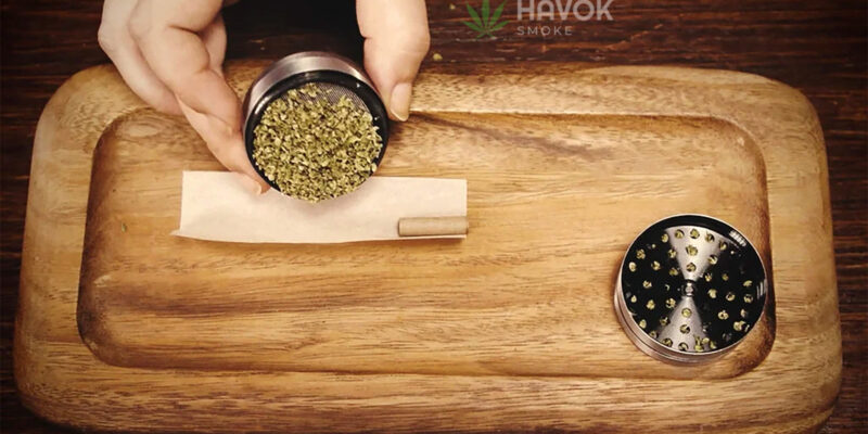 Hand holding a metal weed grinder, pouring freshly ground cannabis onto a rolling paper with a pre-rolled filter tip on a wooden tray. An open grinder with remaining cannabis pieces is placed nearby. HAVOK SMOKE logo is visible in the background.